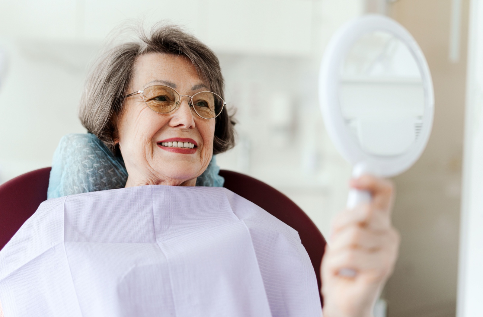 An older adult wearing glasses smiles while looking into a handheld mirror after a dental cleaning.